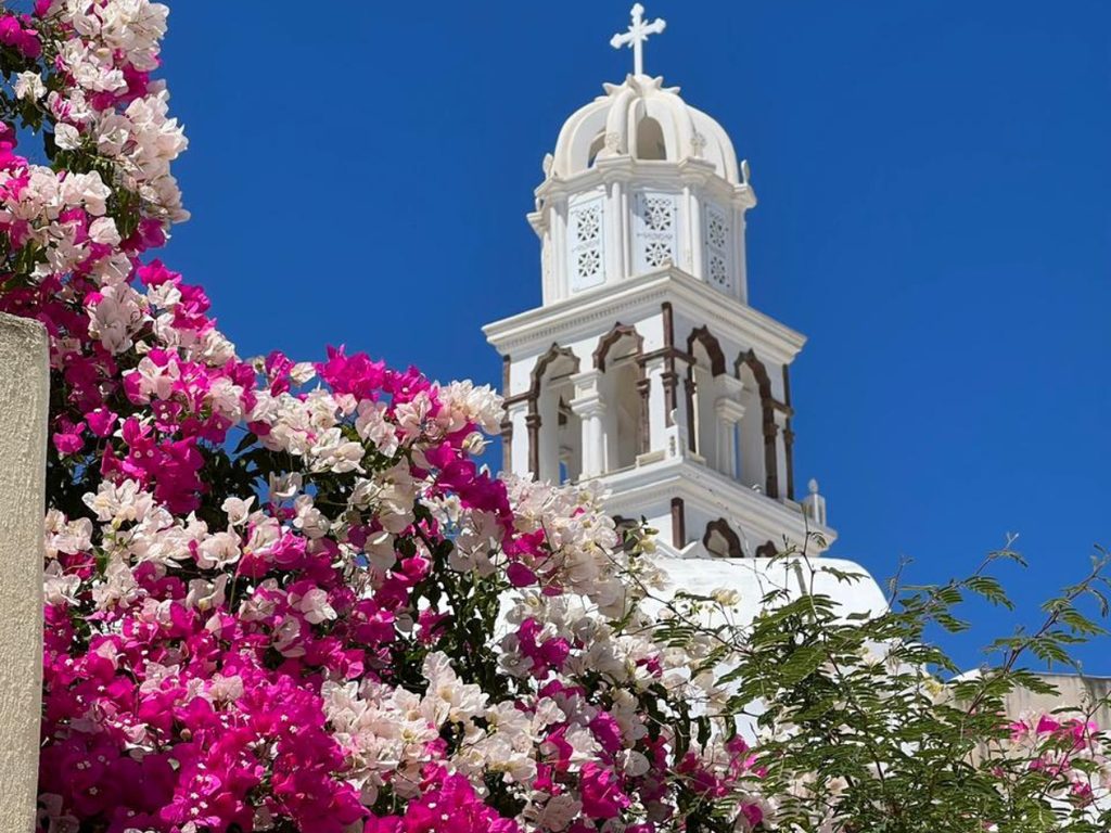 A white Santorinian bell tower framed by pink and white bougainvillea against a deep blue sky