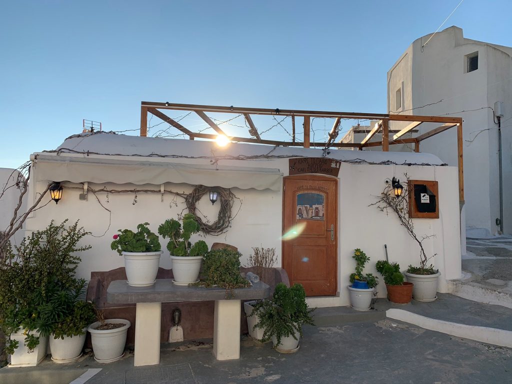 A traditional Santorinian cafe in a village lane at sunset, with potted geraniums and a vine pergola